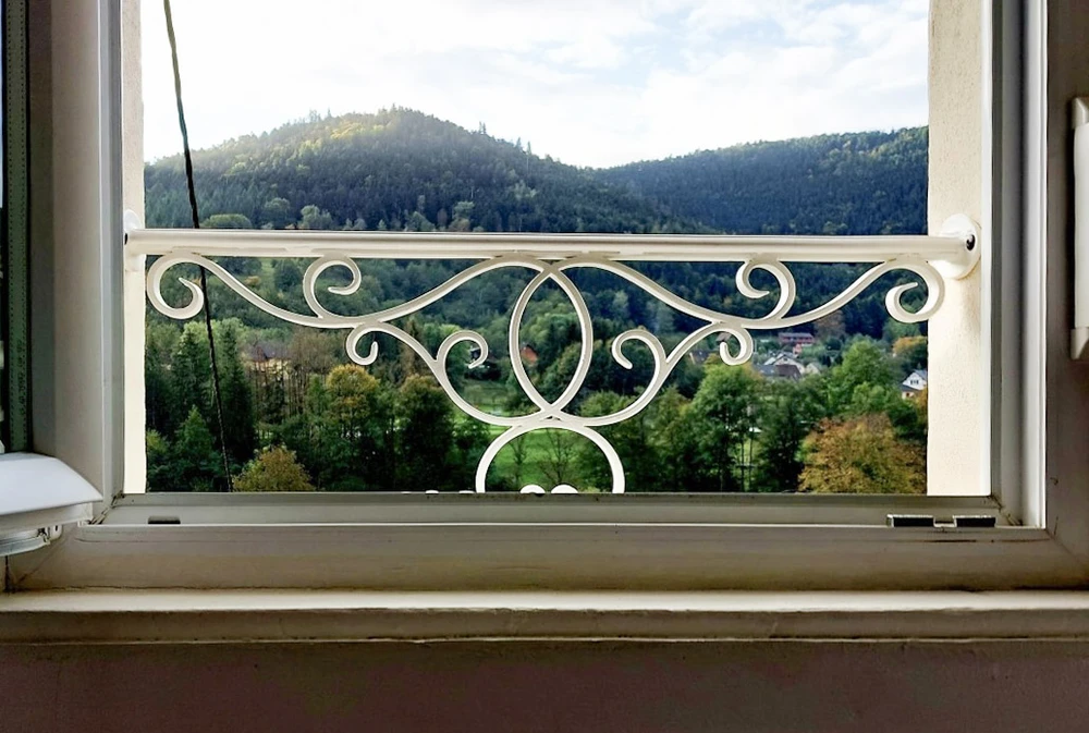 Vue depuis l'intérieur d'une habitation à travers une fenêtre ouverte avec garde-corps décoratif en fer forgé blanc à motifs ornementaux